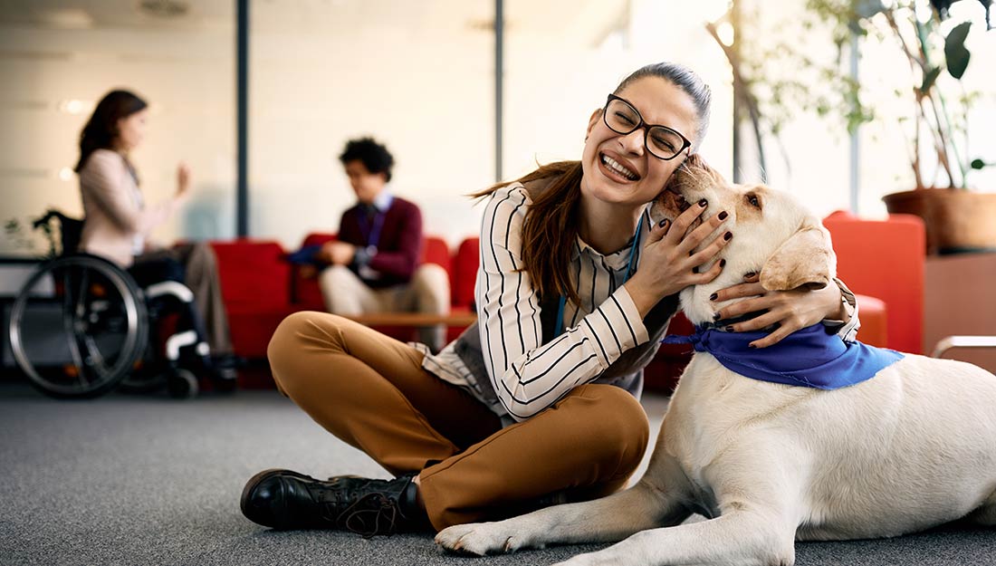 a woman sits on the floor in a workspace, hugging a white dog. people can be seen meeting in the background
