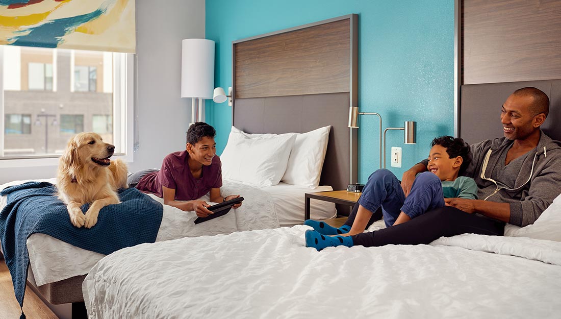 A family sits on beds in a Tru by Hilton hotel. They have a golden retriever with them who resting happily.