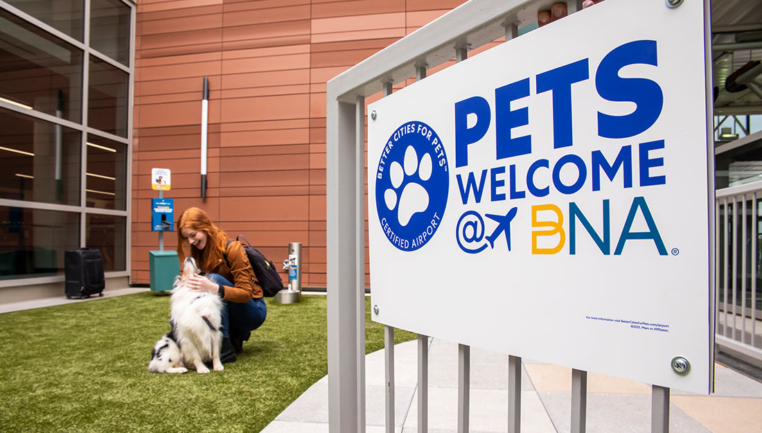 woman with dog at Nashville International Airport (BNA) dog relief area|woman walking with dog in on leash in airport|dog standing in airport waiting area|man with dog entering airport pet relief area|man with dog entering airport pet relief area